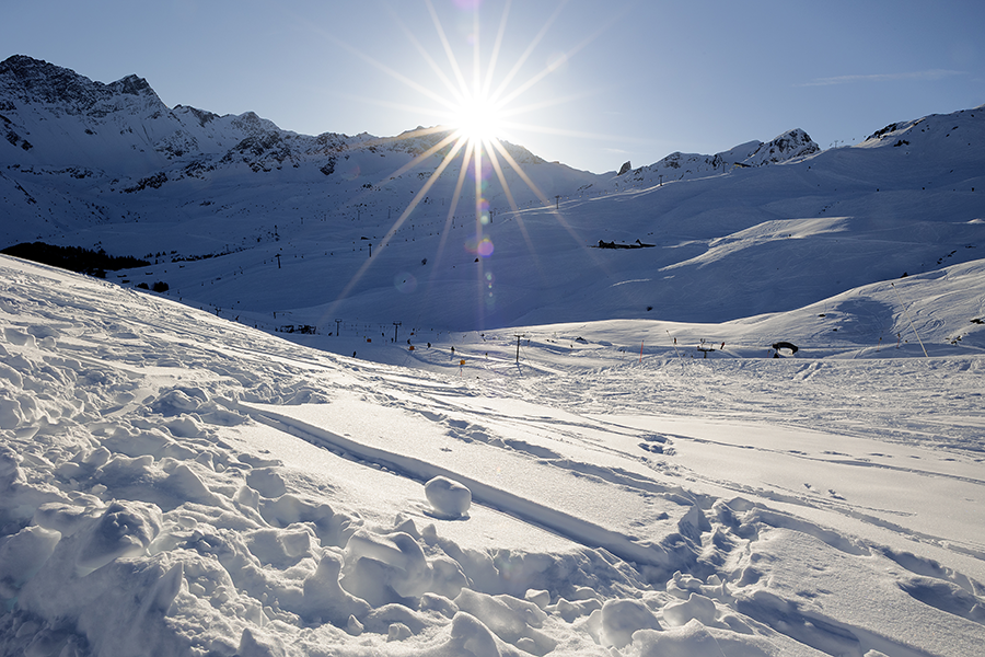 Schneebdeckte Skipiste mit Bergen und Sonne.